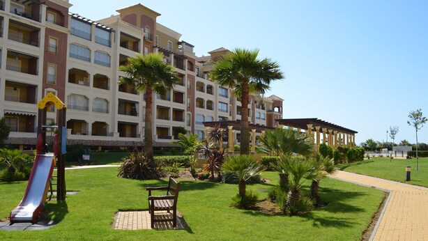 A hotel with multiple balconies, a playground with a slide, and a well-maintained garden with palm trees. - Los Cisnes 13 - Two Bedroom Apartment, Sleeps 5 (Punta del moral)