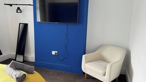 A room with a blue accent wall, a flat-screen TV mounted above a power outlet, a white armchair, and a rolled-up towel on a yellow textured rug. - City Centre-4 Br-sleeps 7-garden-free Parking (Newcastle-upon-Tyne)