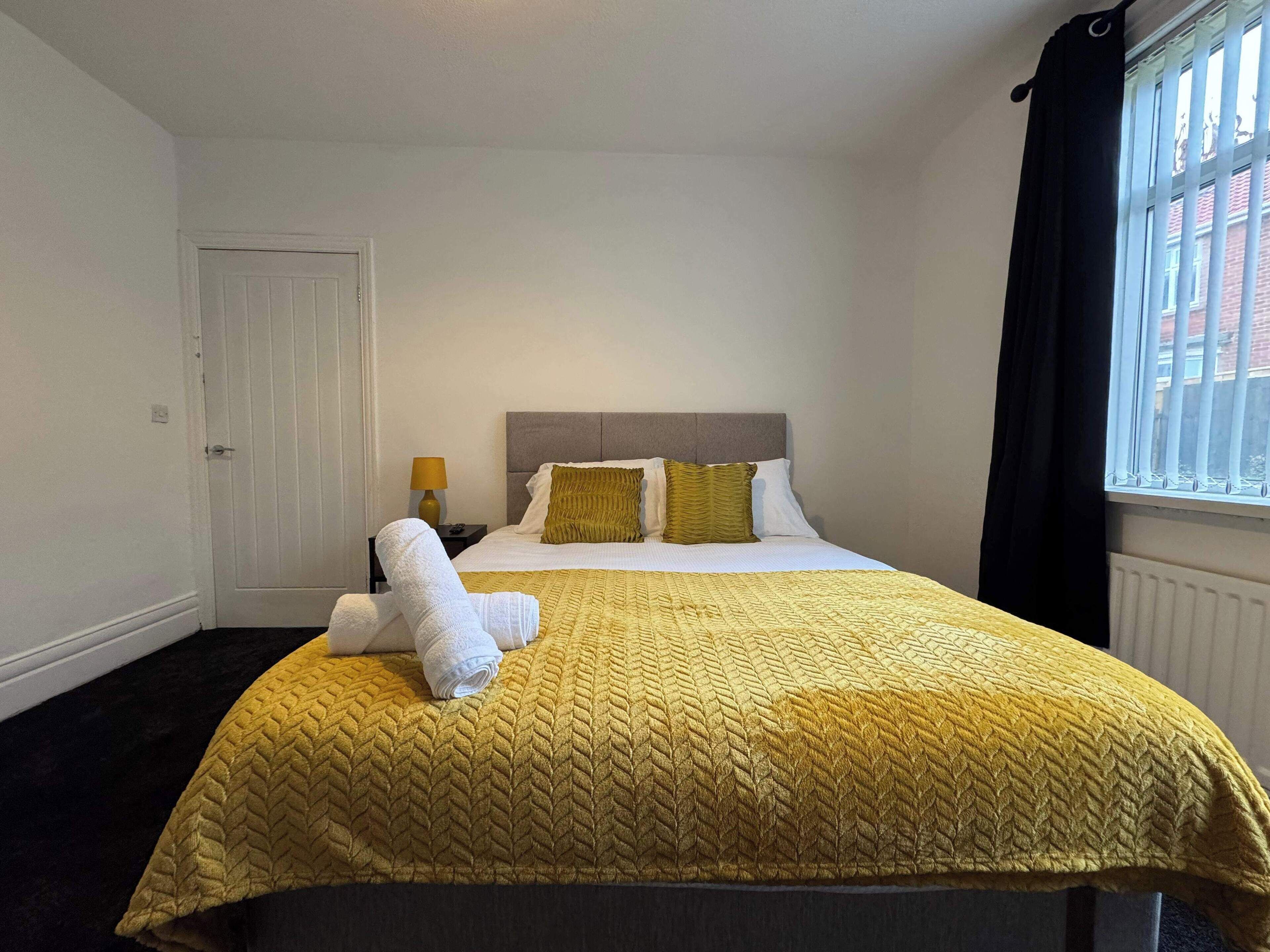 A neatly made bed with a yellow quilt, a white towel, and two green pillows.