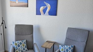 Twee grijze fauteuils met gestreepte kussens, een klein houten bijzettafeltje en twee ingelijste kunstwerken aan een muren met een ruwe textuur.