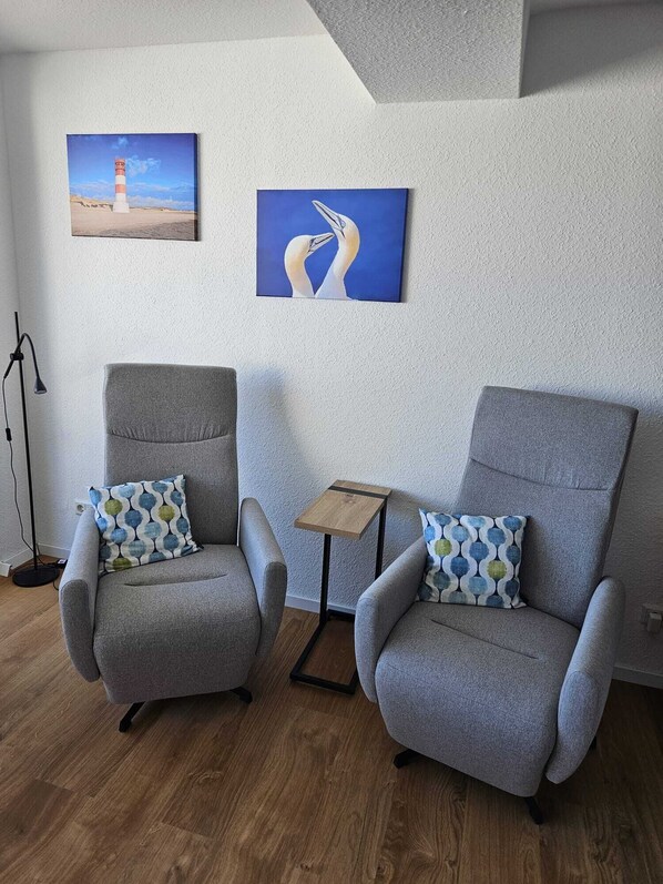 Dos sillones grises con cojines estampados, una mesita de madera y dos cuadros en una pared con textura.