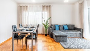 Ein modernes Wohnzimmer mit einem grauen Sofa, schwarzen Sesseln, einem Esstisch für vier Personen und einer großen Pflanze.
