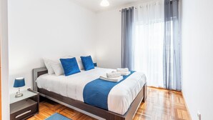 Ein ordentlich bezogenes Bett mit weißer und blauer Bettwäsche, ein Nachttisch mit Lampe und ein Fenster mit transparenten Vorhängen.