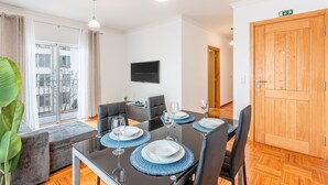 Ein moderner Essbereich mit einem schwarzen Tisch, gedeckt für vier Personen, einer grauen Couch und einem großen Fenster mit Vorhängen.