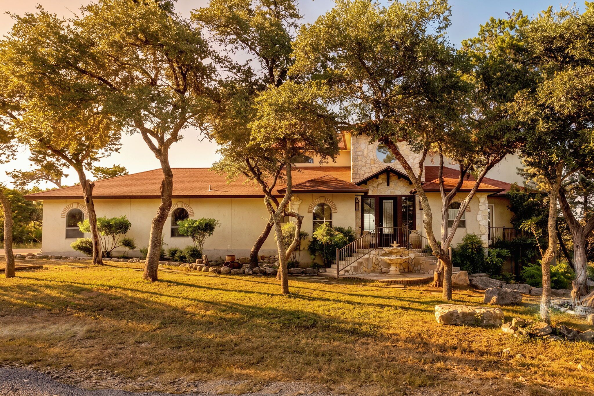 A house with a stone facade, a red roof, and a front yard with trees and a stone fountain.