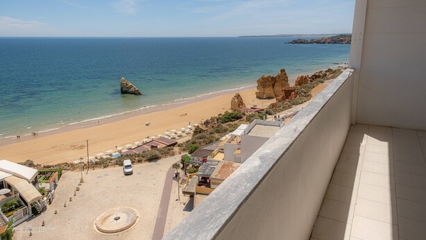 A beach view from a balcony with a clear sky, a sandy beach, and rocky formations. - Apartament Sea View 8E (Portimão)