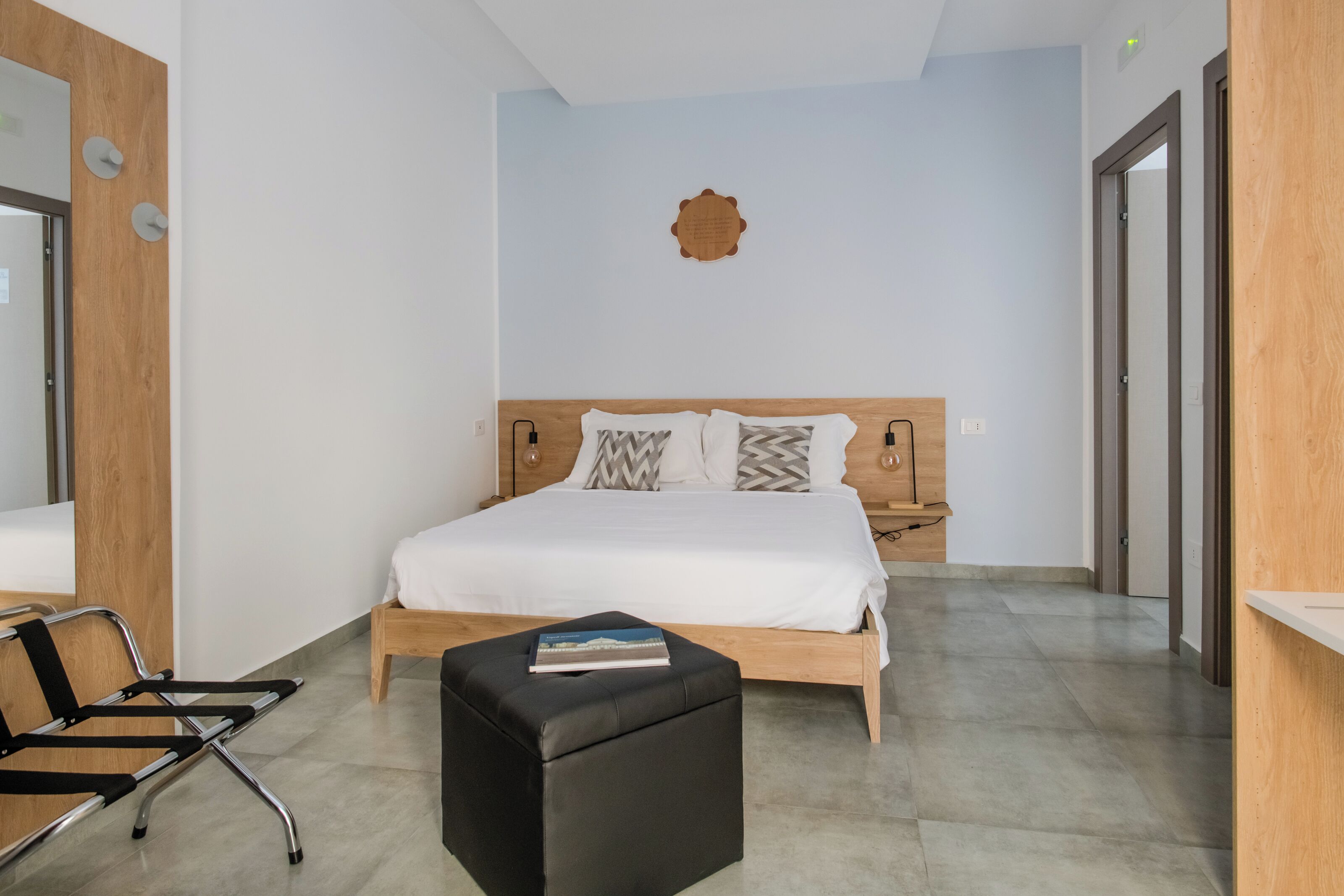 A modern hotel room with a wooden headboard, a large bed, a black ottoman, a wooden bench, and a small table.