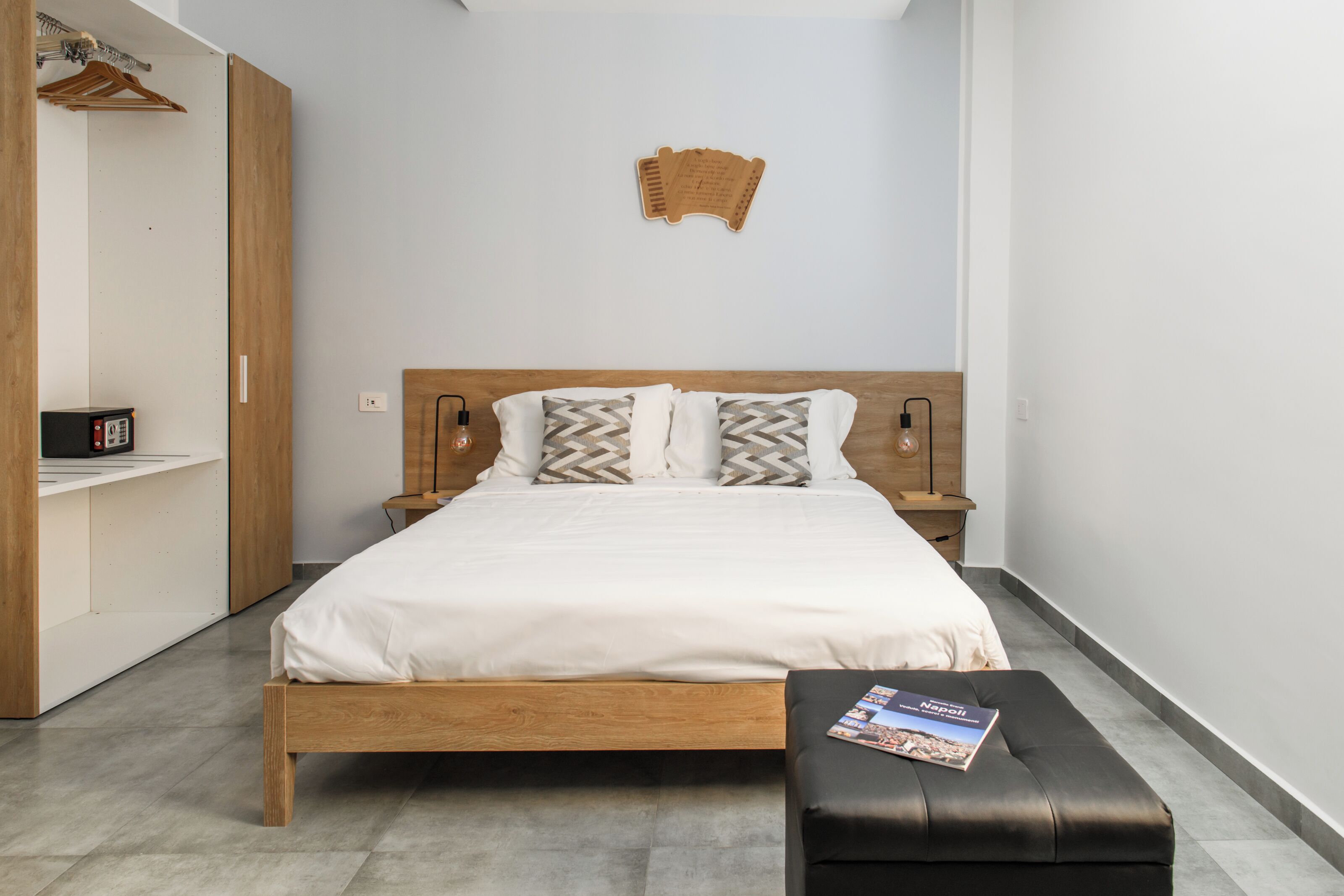 A neatly arranged bedroom with a wooden bed, a bedside table, a nightstand, a wardrobe, and a wall-mounted light fixture.