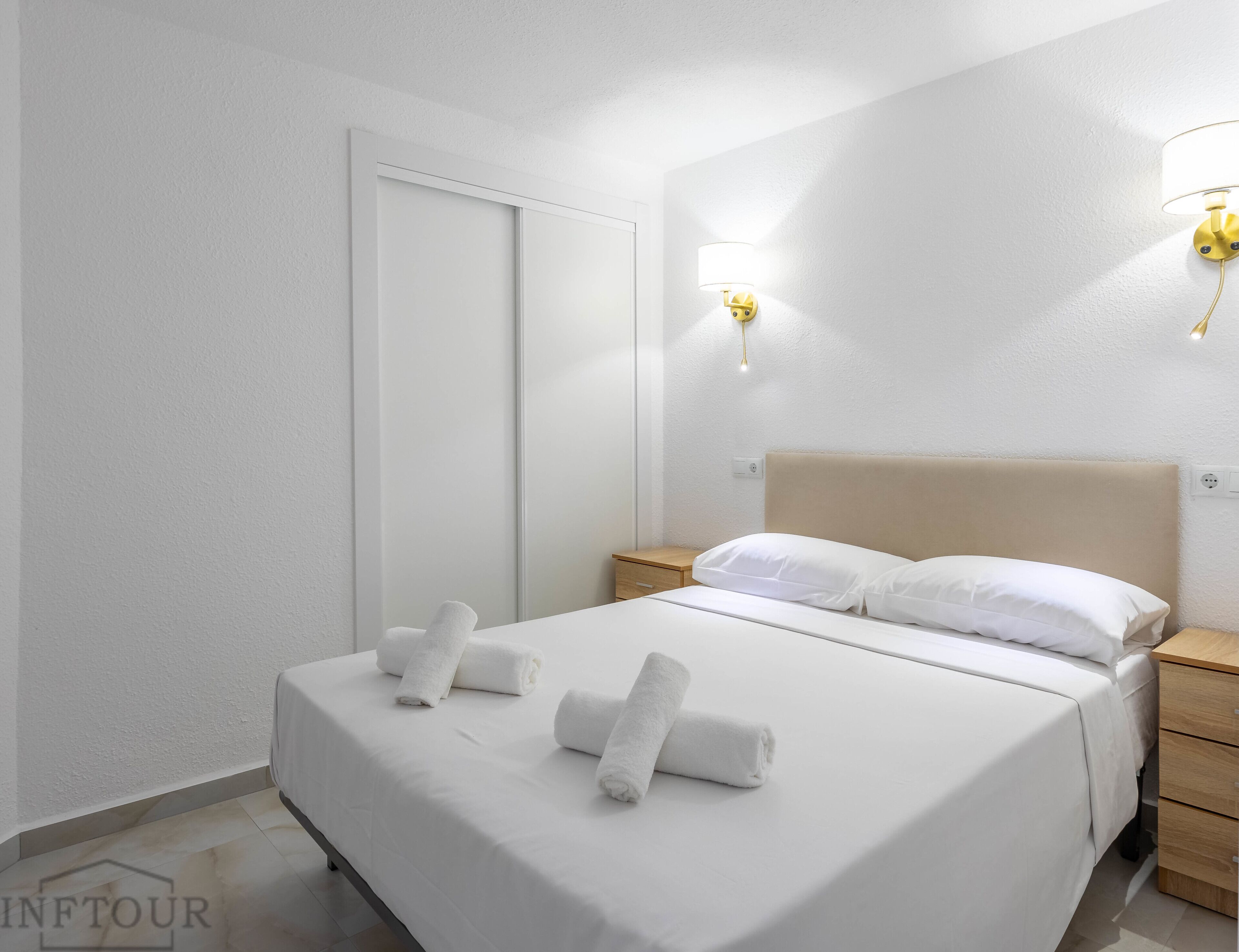 A neatly made bed with white linens and towels, a wooden nightstand, and a wall-mounted lamp.