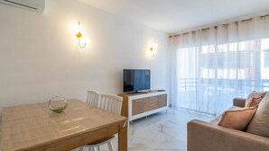 A modern living room with a wooden dining table, white chairs, a TV on a wooden cabinet, and a balcony with sheer curtains.