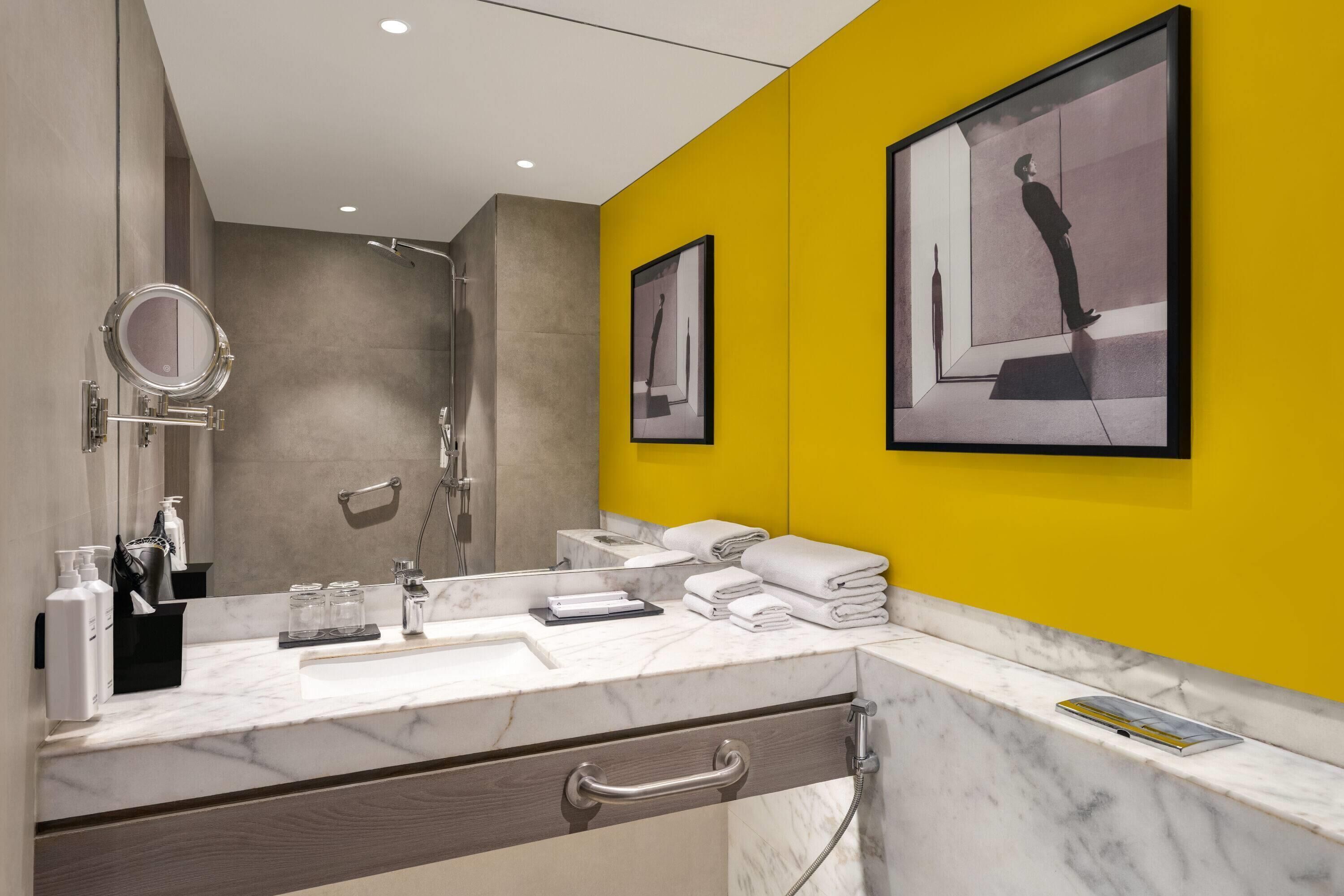 Un baño moderno con tocador de mármol, un espejo grande, una pared de acento amarilla y una obra de arte enmarcada.