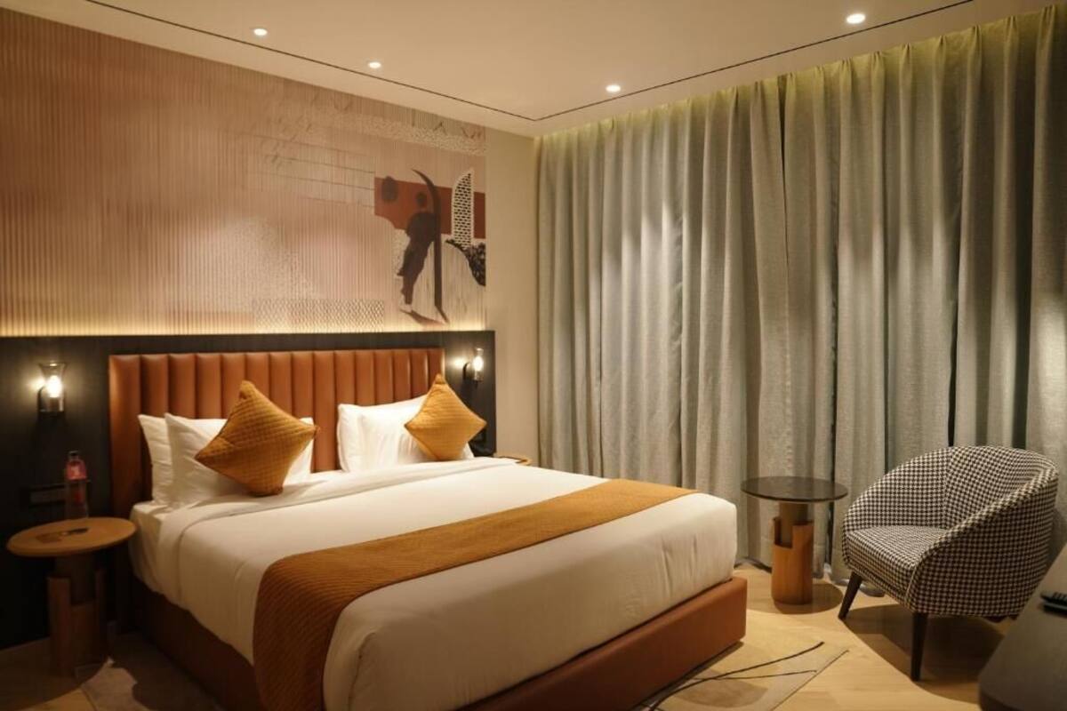 A modern hotel room with a large bed, a headboard with a textured design, a round side table, a chair, and a large window with curtains.
