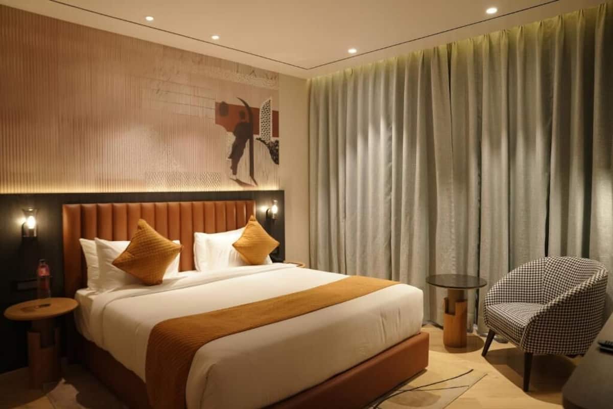 Une chambre d’hôtel moderne avec un grand lit, une tête de lit au motif texturé, une table d’appoint ronde, une chaise et une grande fenêtre avec des rideaux.