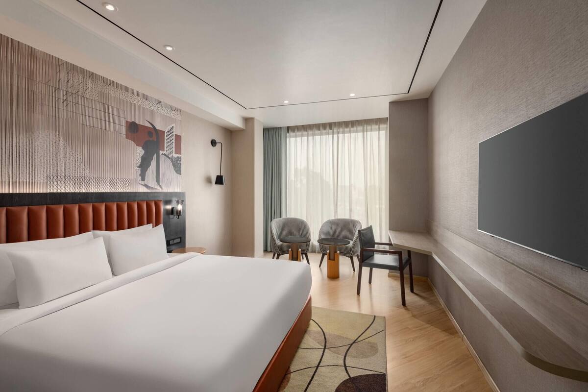 A modern hotel room with a large bed, two chairs, a small table, a TV, and a decorative wall piece.