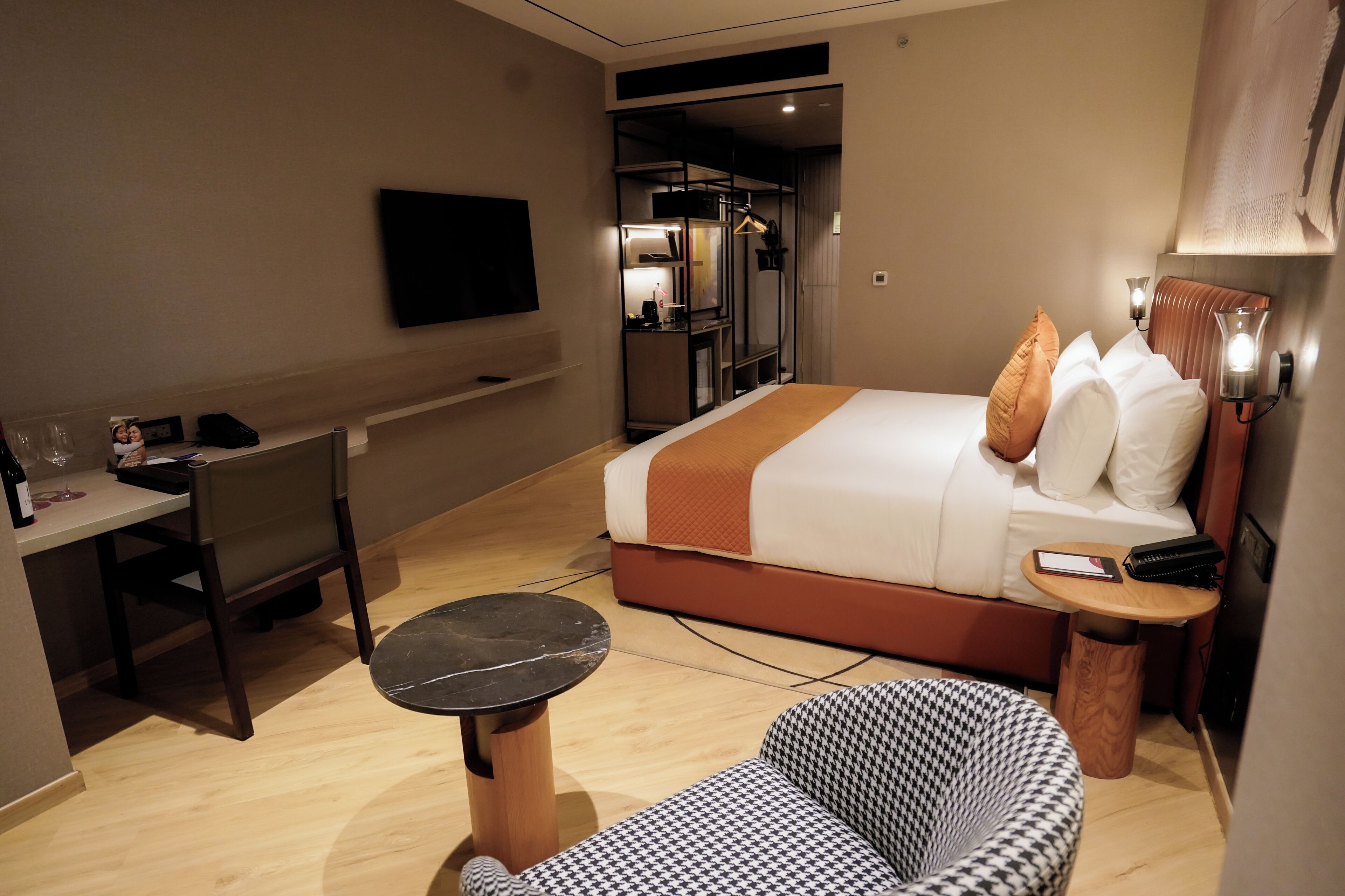 A hotel room with a bed, a desk, a chair, a small table, and a TV mounted on the wall.