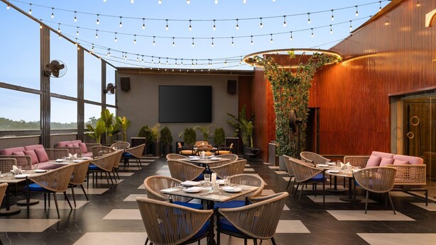 A restaurant with round tables and wicker chairs, string lights overhead, and a large screen on the wall. - Ramada Encore By Wyndham Chandigarh Zirakpur (Zirakpur)