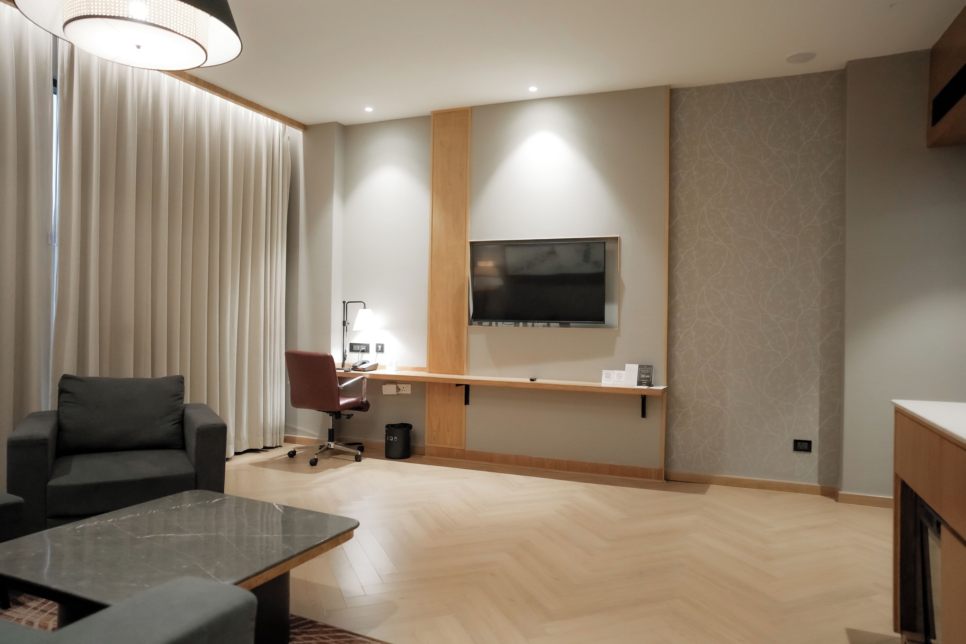 Ein modernes Hotelzimmer mit Wand-Fernseher, einer Couch, einem Schreibtisch mit Stuhl und einem Parkettboden mit Fischgrätenmuster.