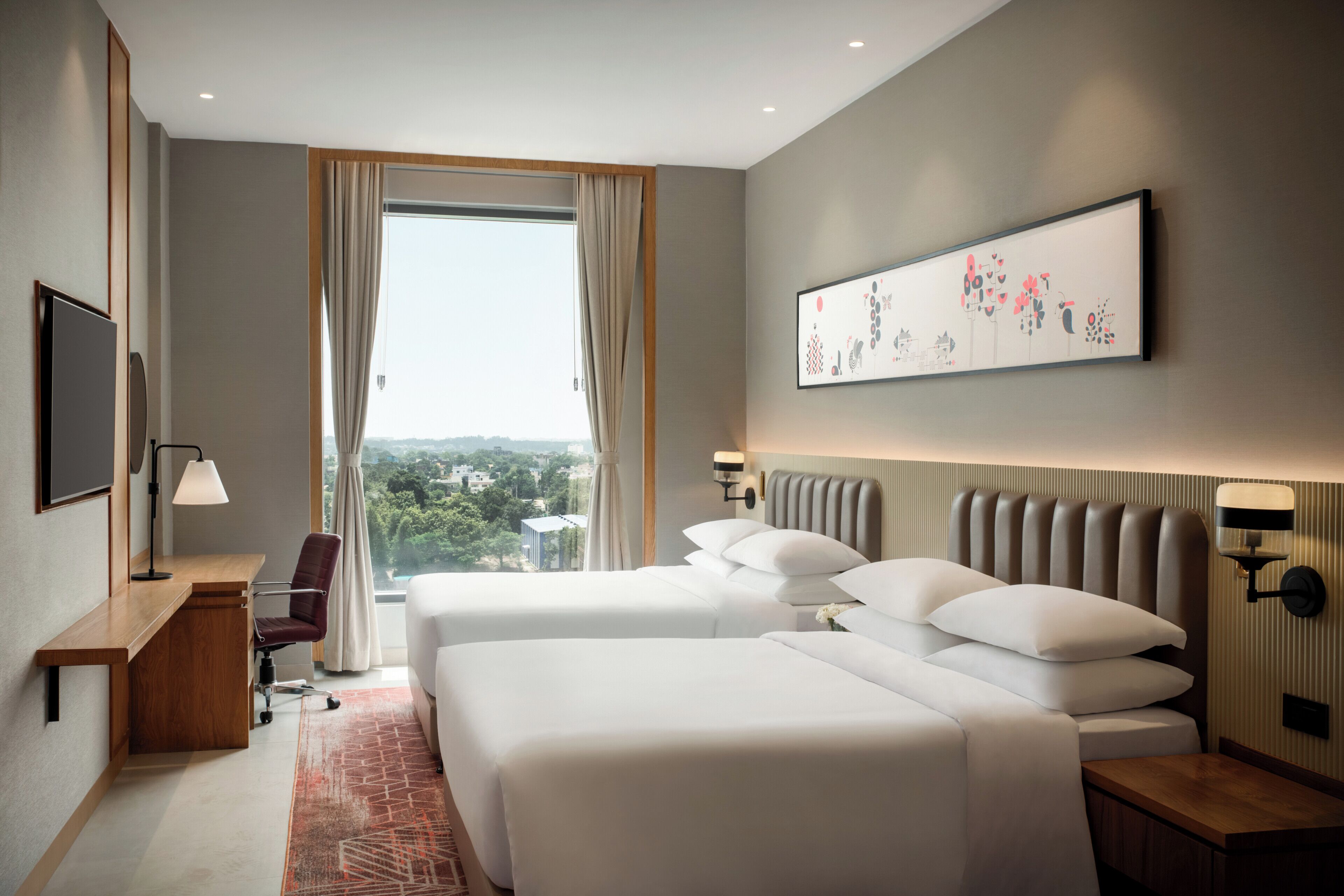 A hotel room with two beds, a desk, a chair, a TV, a window with curtains, and a wall-mounted artwork.