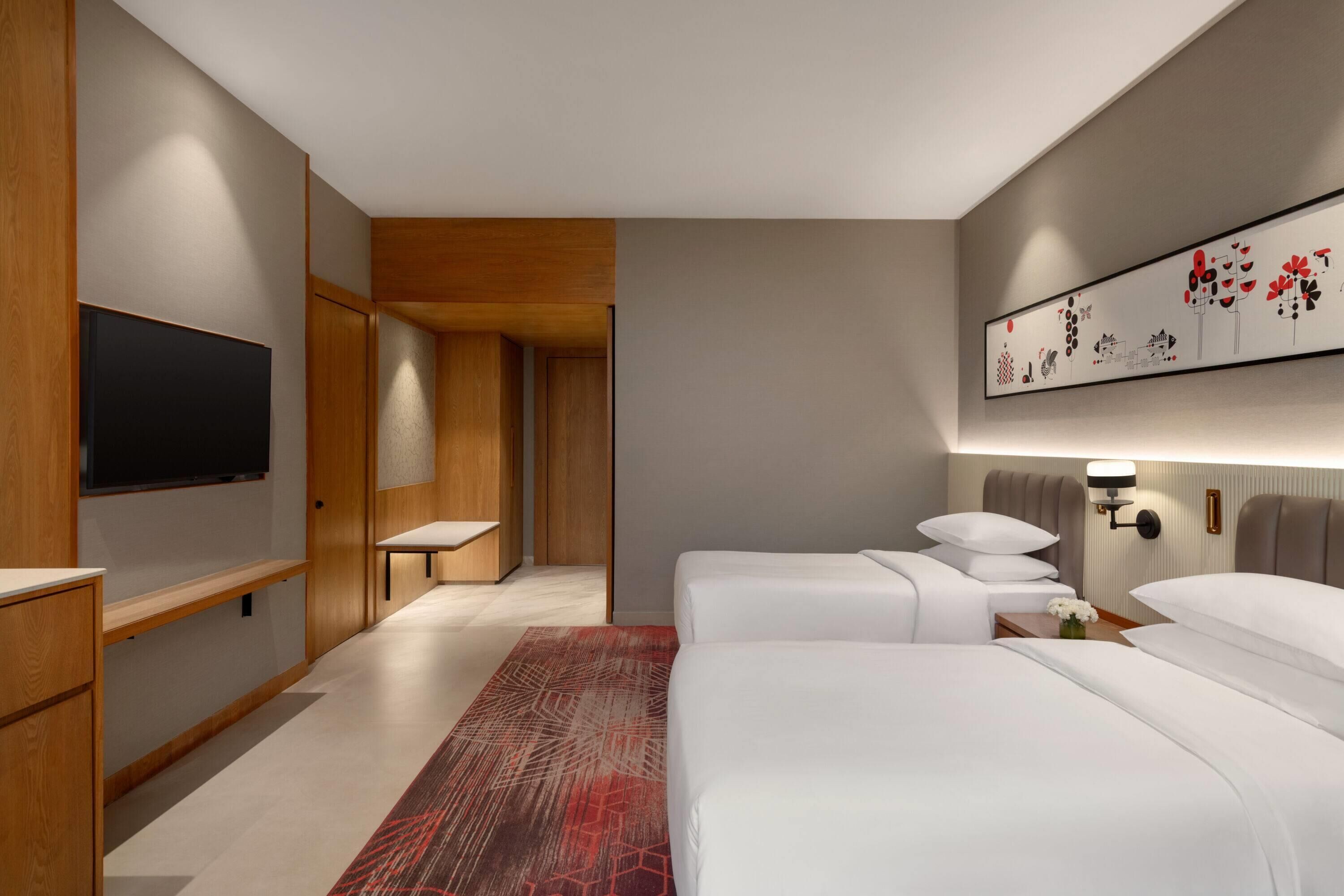A modern hotel room with a flat-screen TV, two beds, a red patterned rug, and a wall-mounted artwork.