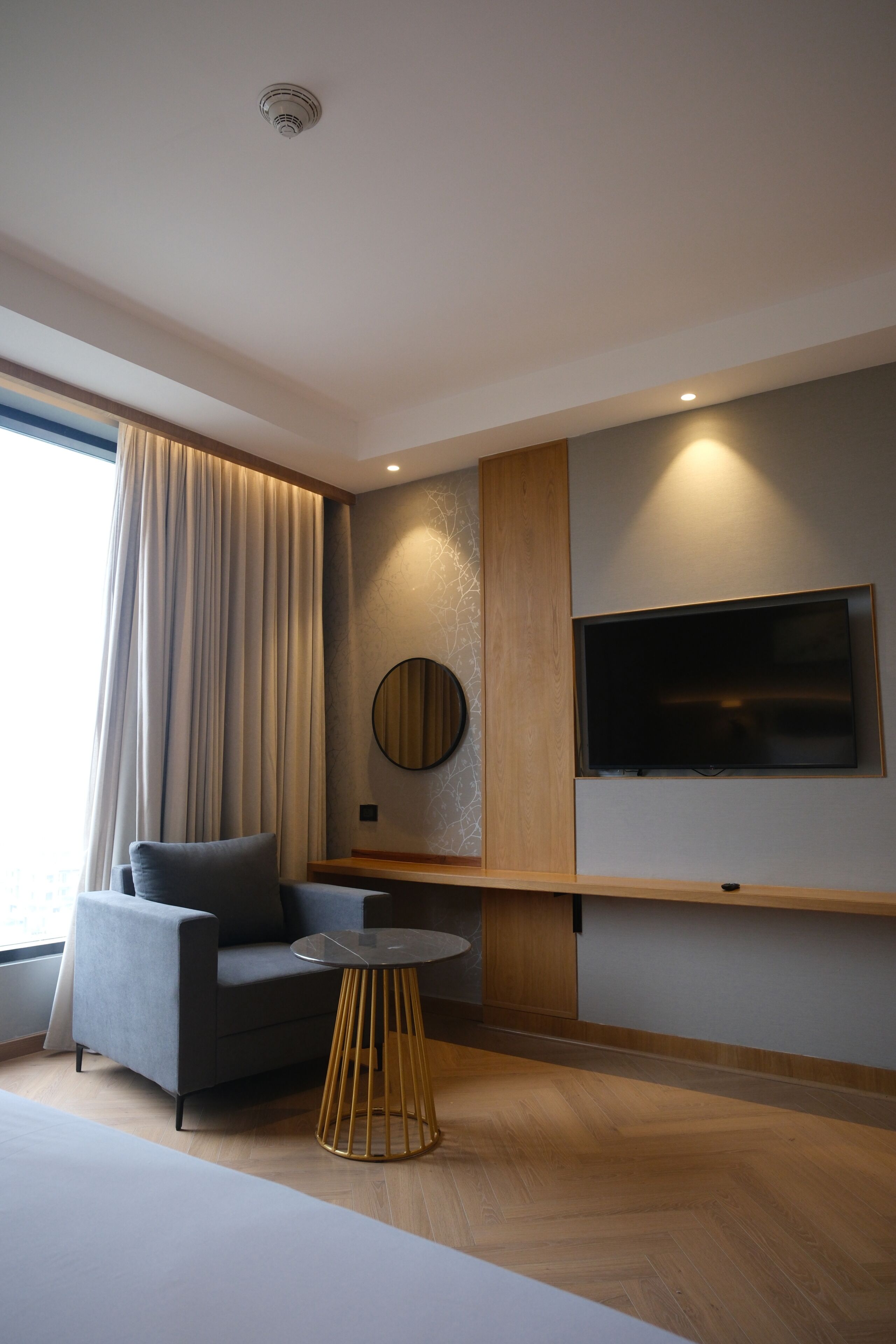 A modern hotel room with a sofa, a round table, a mounted TV, and a large window with curtains.