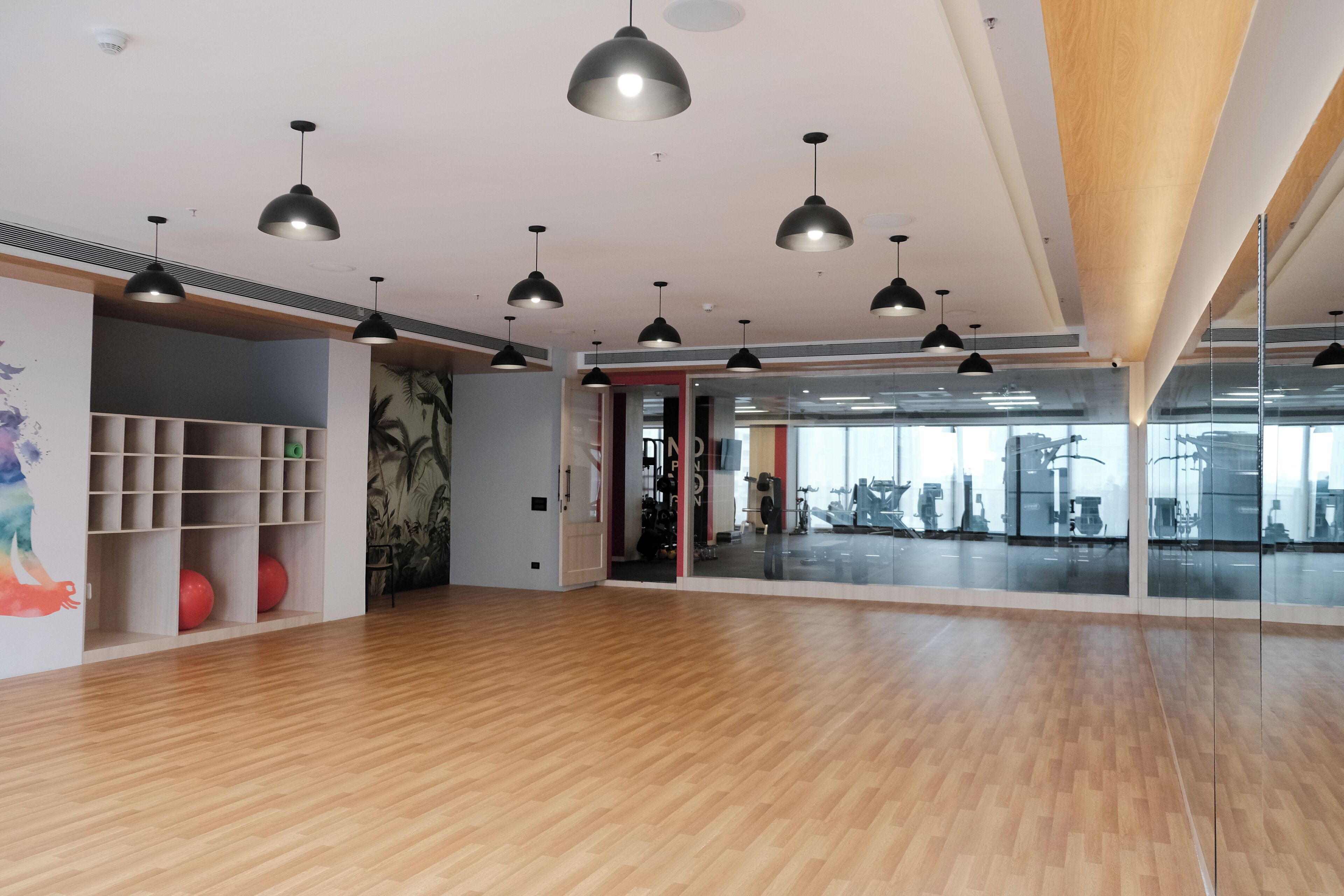 A spacious room with wooden flooring, black pendant lighting, a mirrored wall, and a door leading to a gym area.