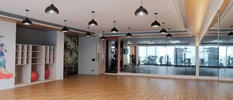 A spacious room with wooden flooring, black pendant lighting, a mirrored wall, and a door leading to a gym area.