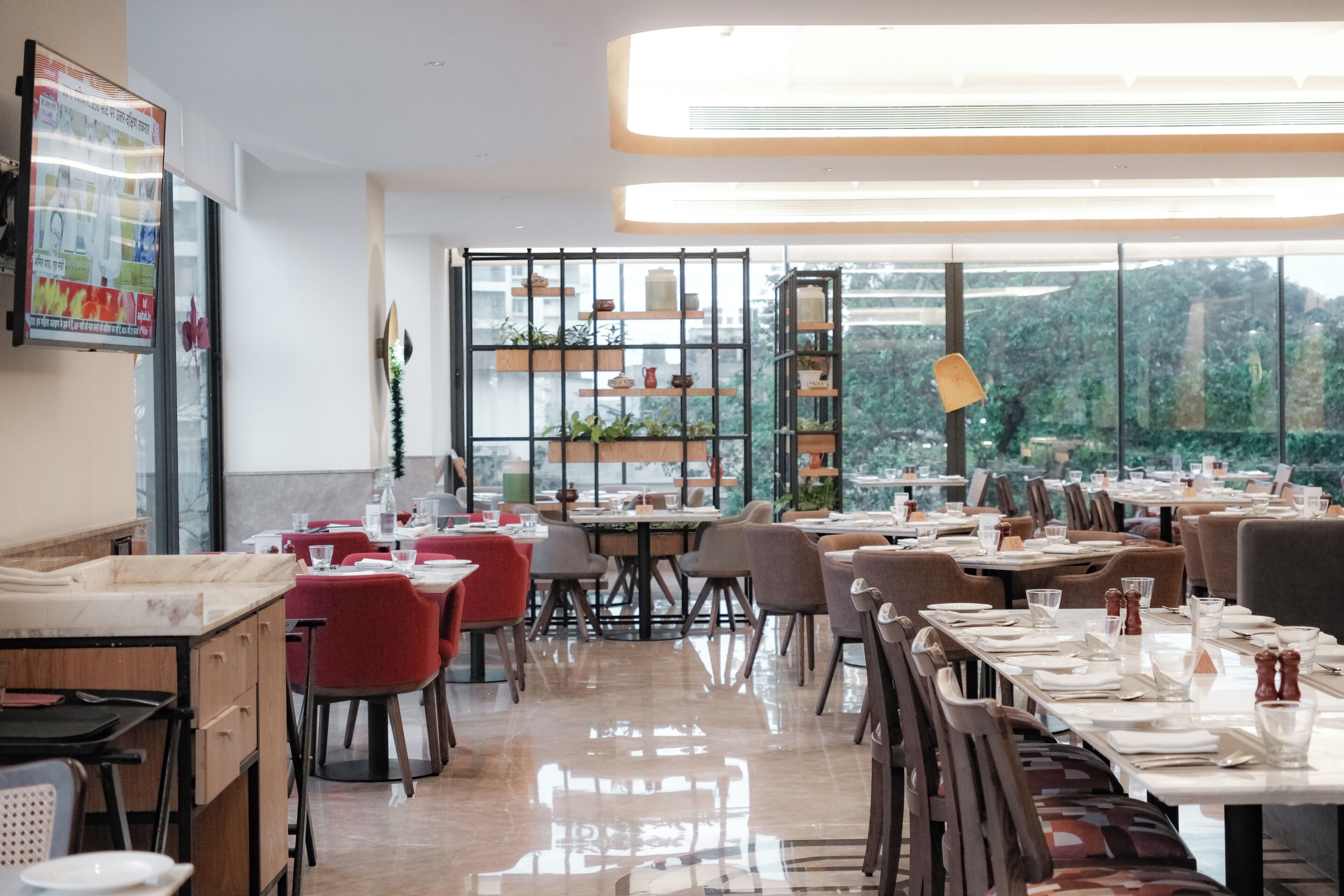 A restaurant with tables set for dining, a large window with a view, and shelves with plants.