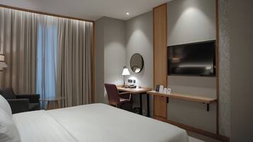 A hotel room with a bed, a desk with a chair, a TV, and a window with curtains.