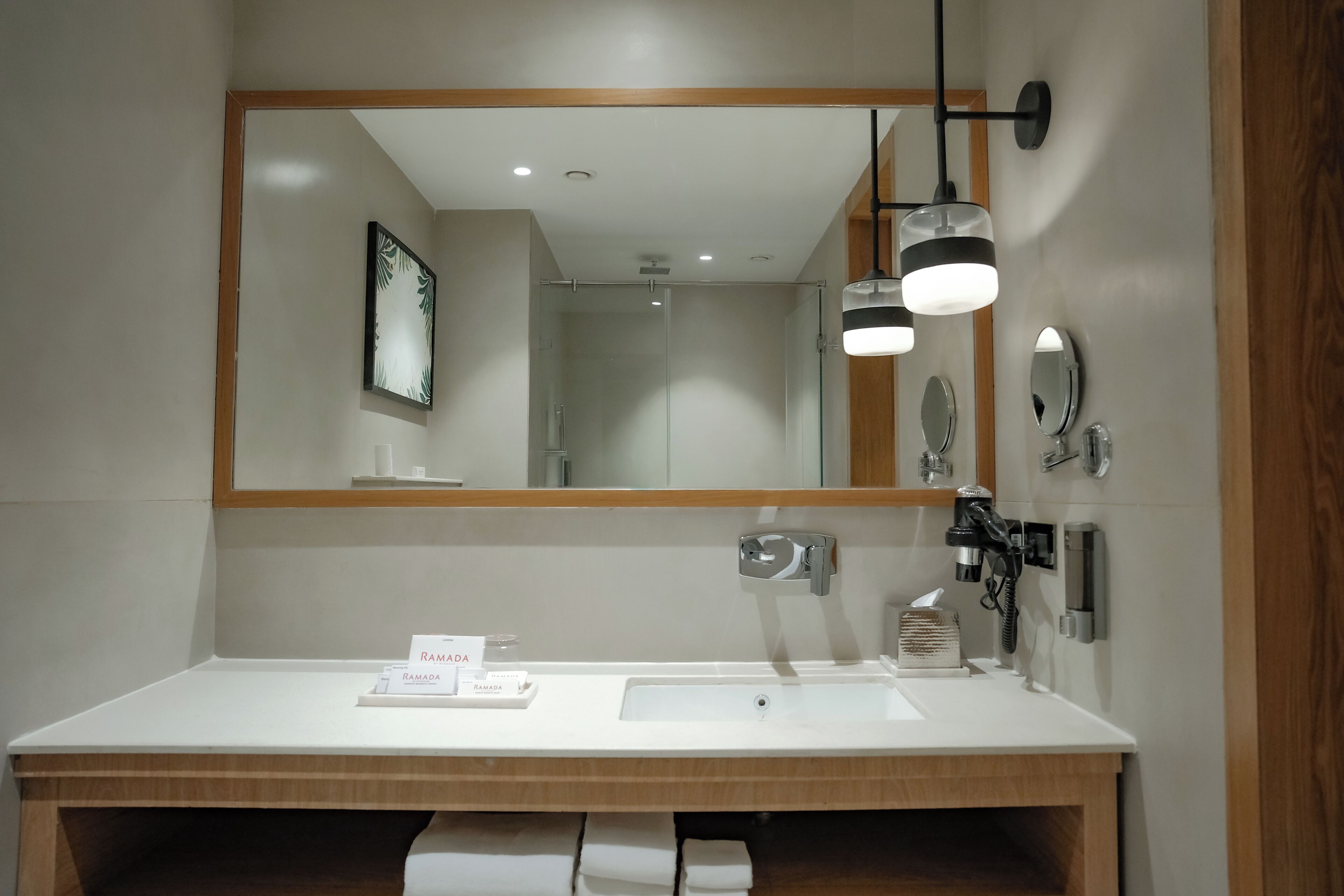 Un baño moderno con un espejo grande, un lavabo blanco, un perchero y un cartel de 'RAMADA'.