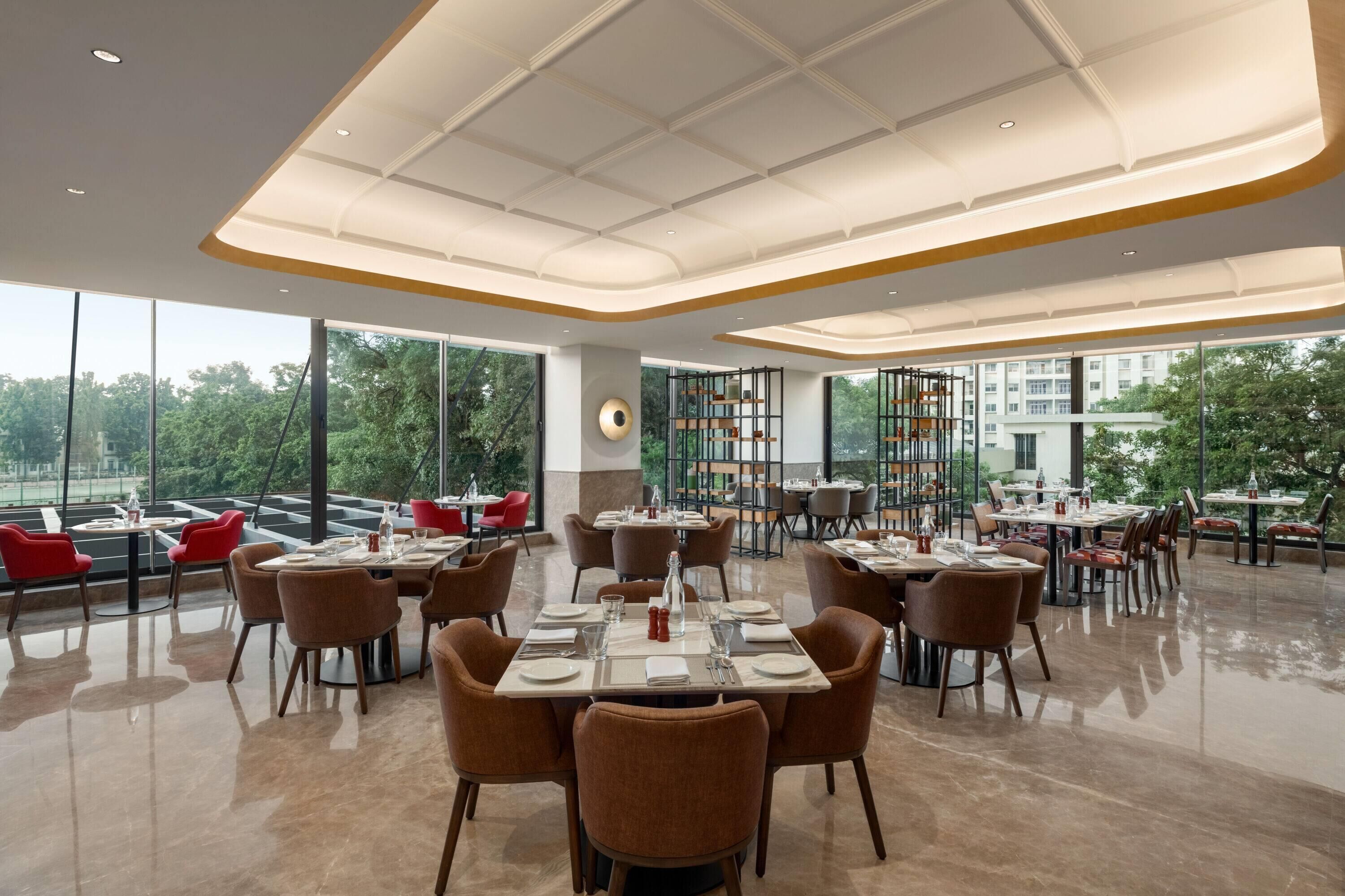 Un restaurant spacieux avec de hauts plafonds et de grandes fenêtres offrant une vue sur la ville. Les tables sont disposées avec soin et accompagnées de chaises, et le sol est ciré.