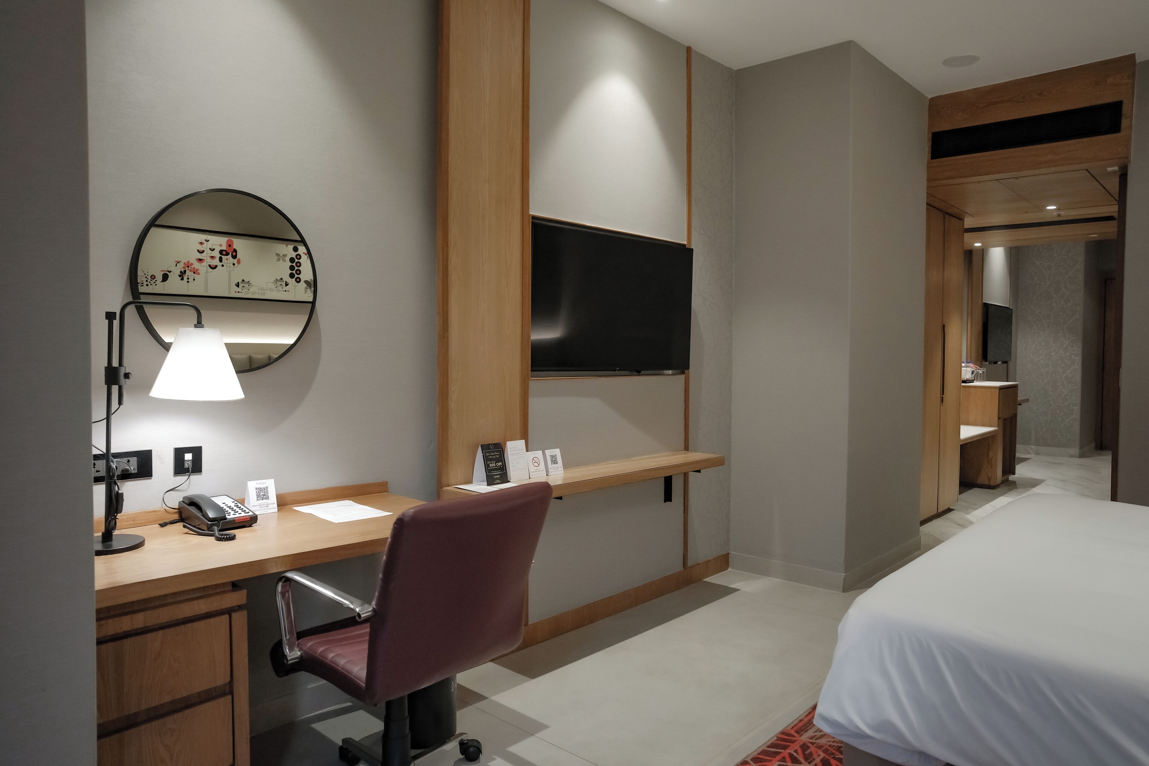A hotel room with a desk, chair, bed, TV, and a mirror with artwork.