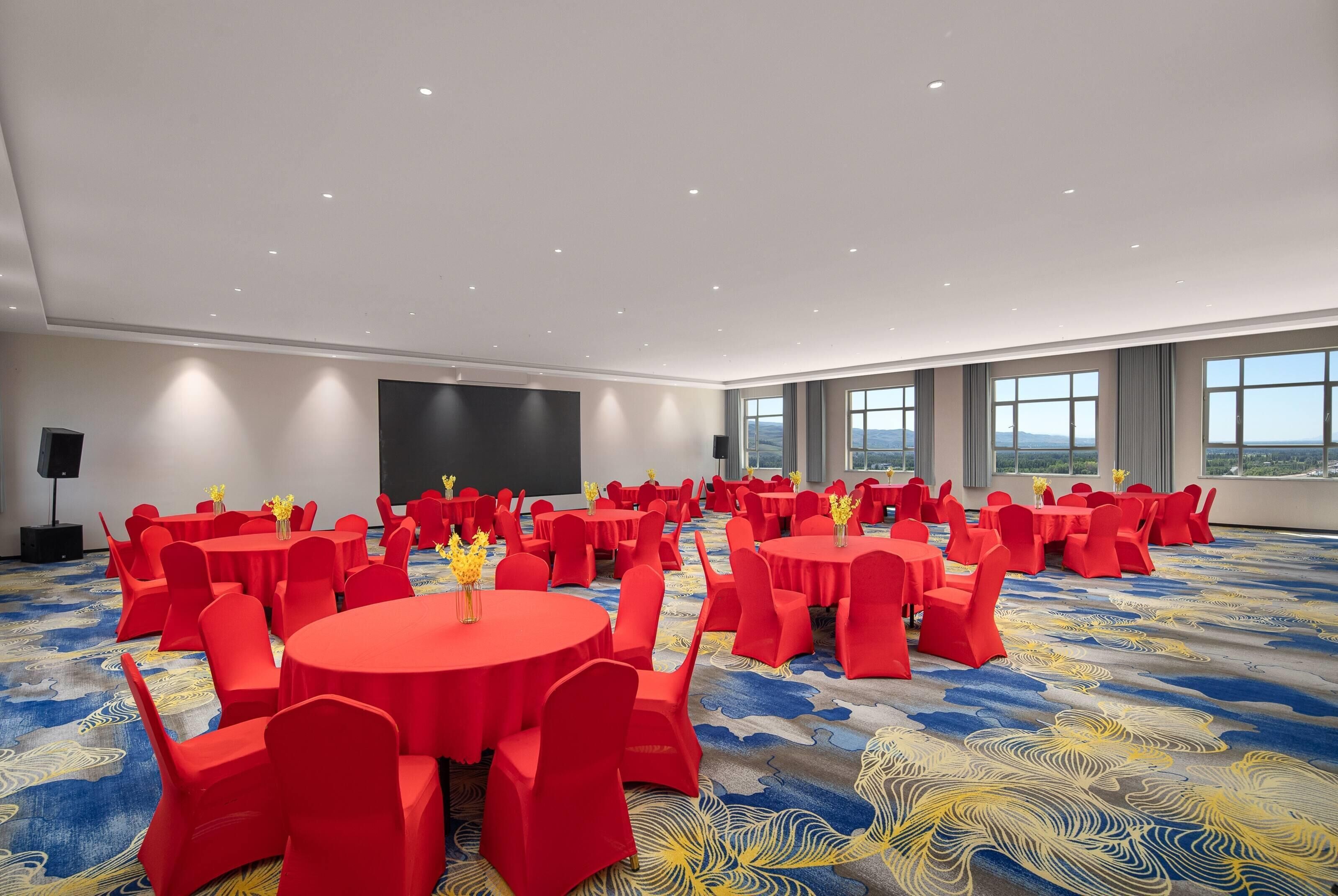 Ein großer Konferenzraum mit mehreren runden Tischen und roten Stühlen, mit einem blau-gelben Teppichboden und großen Fenstern mit Blick auf den Himmel.