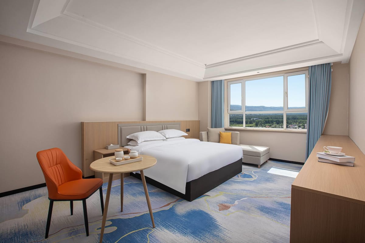 Quarto de hotel com cama, escrivaninha, cadeira, mesinha e janela com vista.