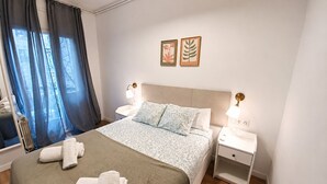 Una cama bien hecha con una colcha estampada, dos mesitas de noche con lámparas, una ventana con cortinas y dos cuadros enmarcados en la pared.