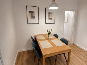Ruokailutilassa on puinen pöytä, siniset tuolit, vihreitä kasveja sisältävä maljakko sekä kaksi seinälle ripustettua kehystettyä kuvaa.
