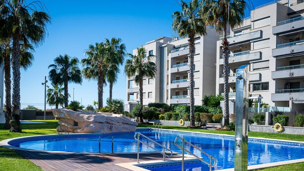 A swimming pool with a wooden deck, palm trees, and a modern building in the background. - Peaceful ground floor with private garden and terrace (Torredembarra)