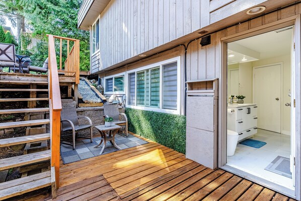 Une terrasse en bois avec des meubles d’extérieur, un escalier et une vue sur une salle de bain dotée d’une porte et de meubles blancs.