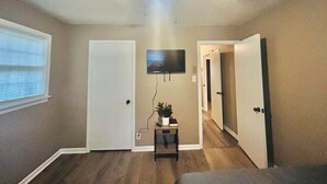 A room with a ceiling fan, a mounted TV, a small table with a plant, and a door leading to another room.