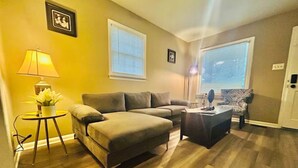 A living room with a sectional sofa, a small round table with a lamp, a patterned armchair, and a large window with blinds.