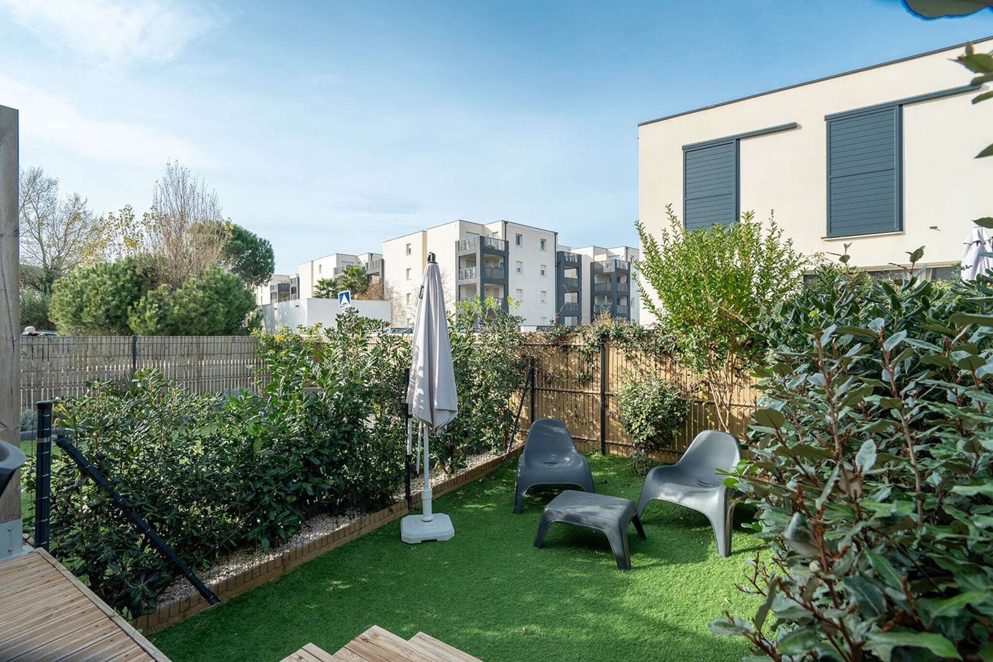 A backyard with artificial grass, a wooden deck, and a patio area with plastic chairs and an umbrella.