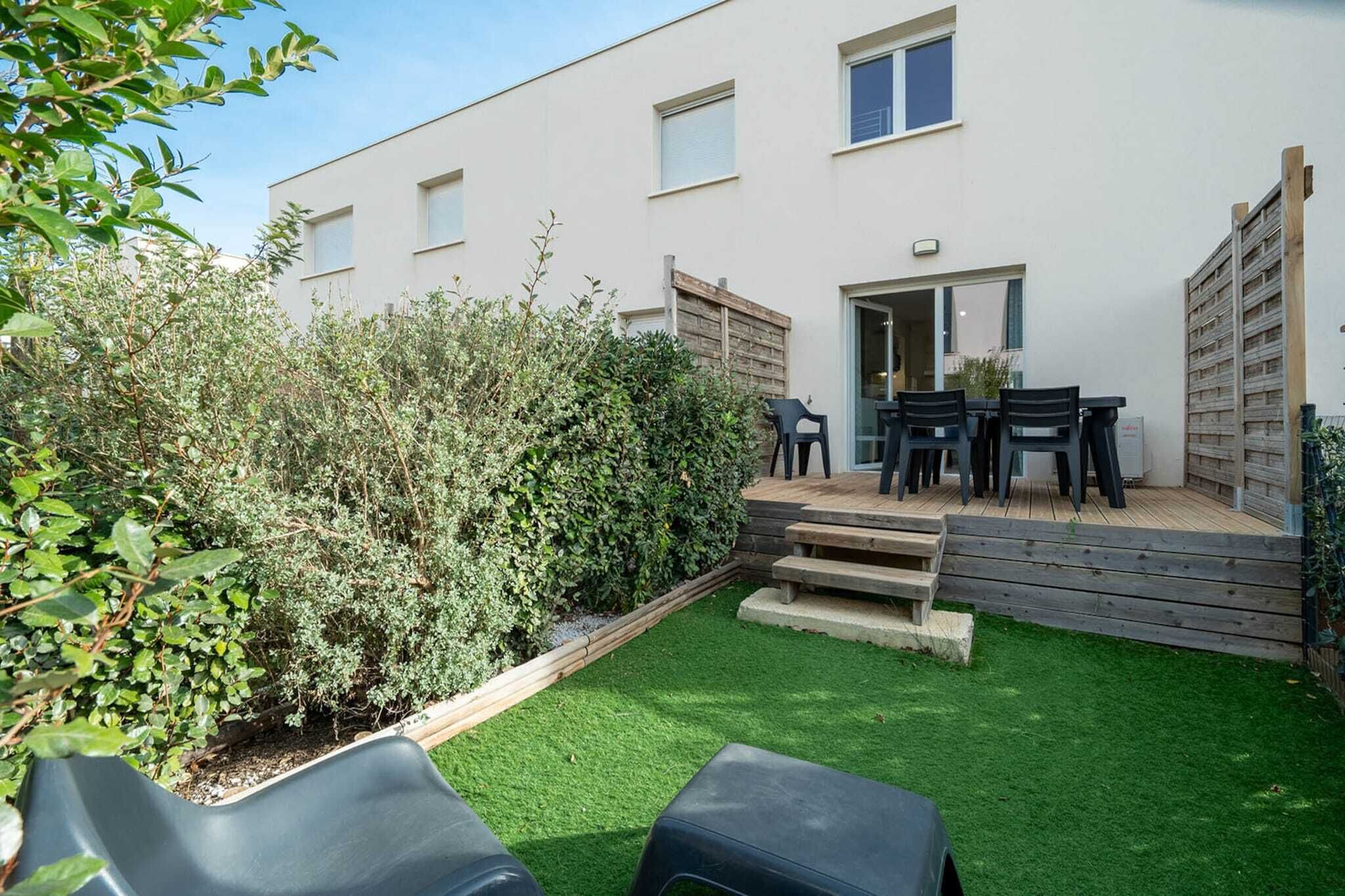 A modern building with a flat roof, a small deck with a table and chairs, and a well-maintained garden with green grass and bushes.
