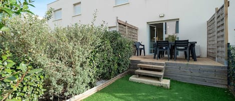 A modern building with a flat roof, a small deck with a table and chairs, and a well-maintained garden with green grass and bushes.