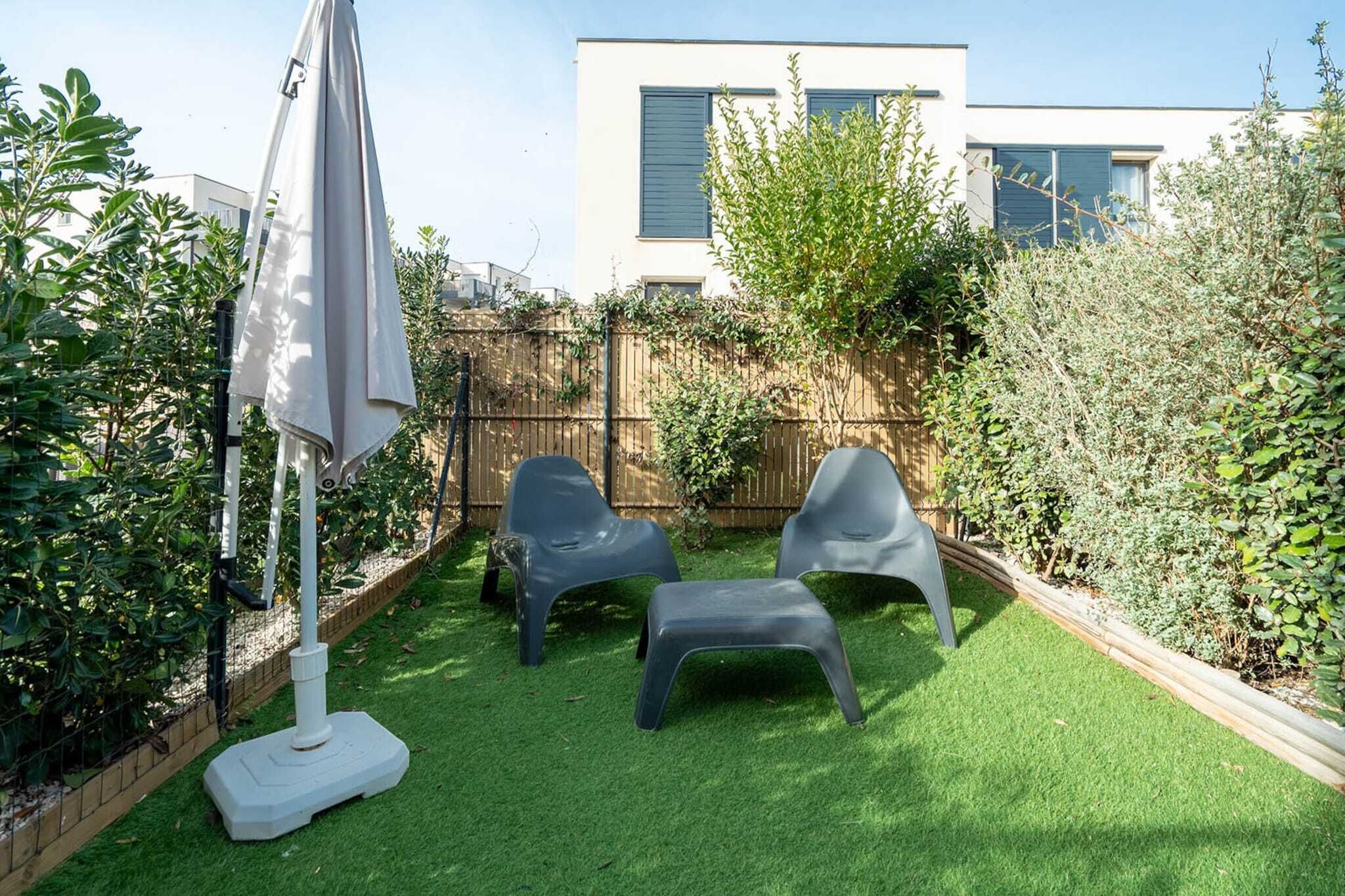 Een achtertuin met kunstgras, twee plastic stoelen, een witte parasol en een houten schutting.