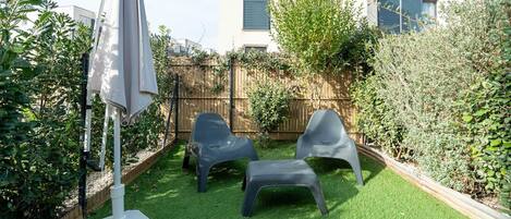 Een achtertuin met kunstgras, twee plastic stoelen, een witte parasol en een houten schutting.