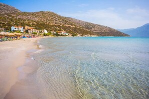 A coastal beach with clear turquoise water, a sandy shore, and buildings on a hillside.