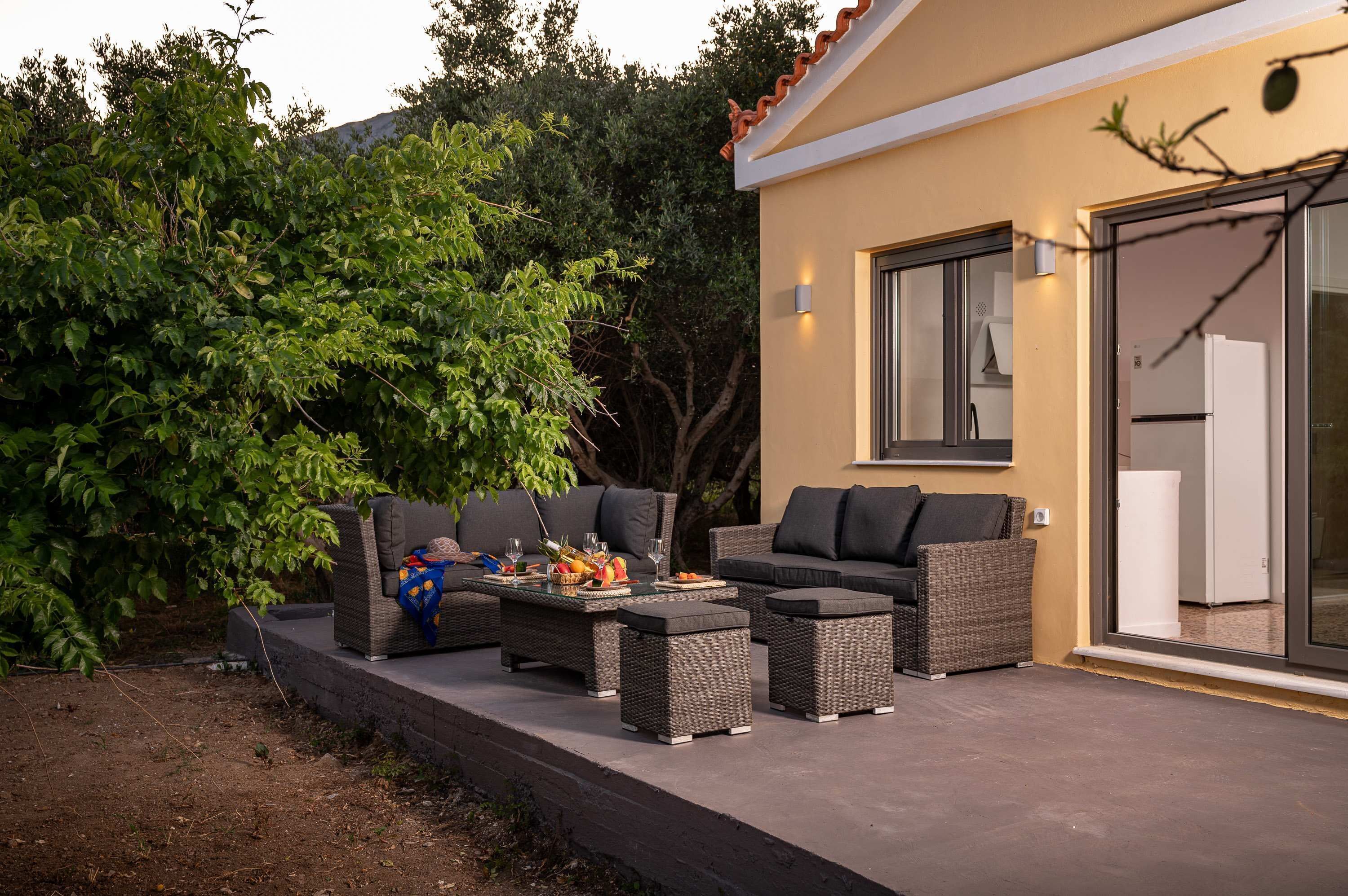 An outdoor patio with wicker furniture, a dining table with fruit and drinks, and a sliding glass door leading to a kitchen.
