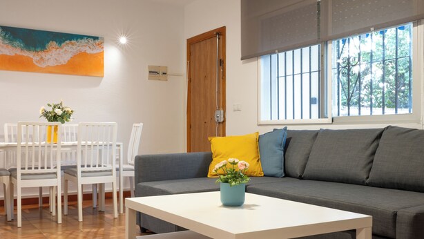 Ein Wohnzimmer mit grauem Sofa, einem weißen Couchtisch und einem Esstisch mit weißen Stühlen. Ein Fenster mit Gitter und ein Bild an der Wand sind ebenfalls zu sehen.