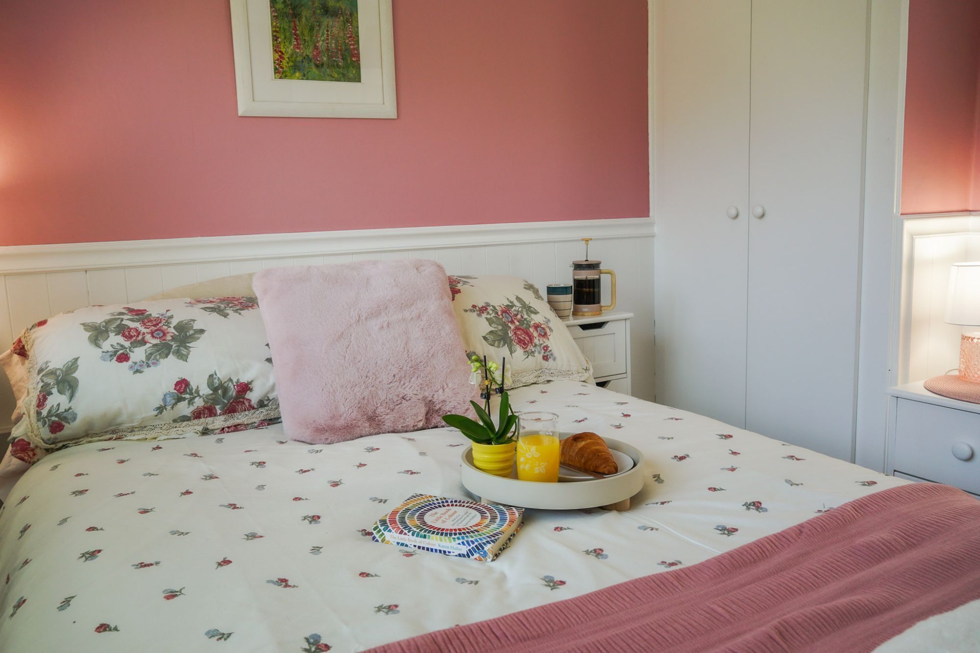 En snyggt bäddad säng med blommigt sänglinne, en rosa pläd och ett nattduksbord med en kaffekanna och en bok.