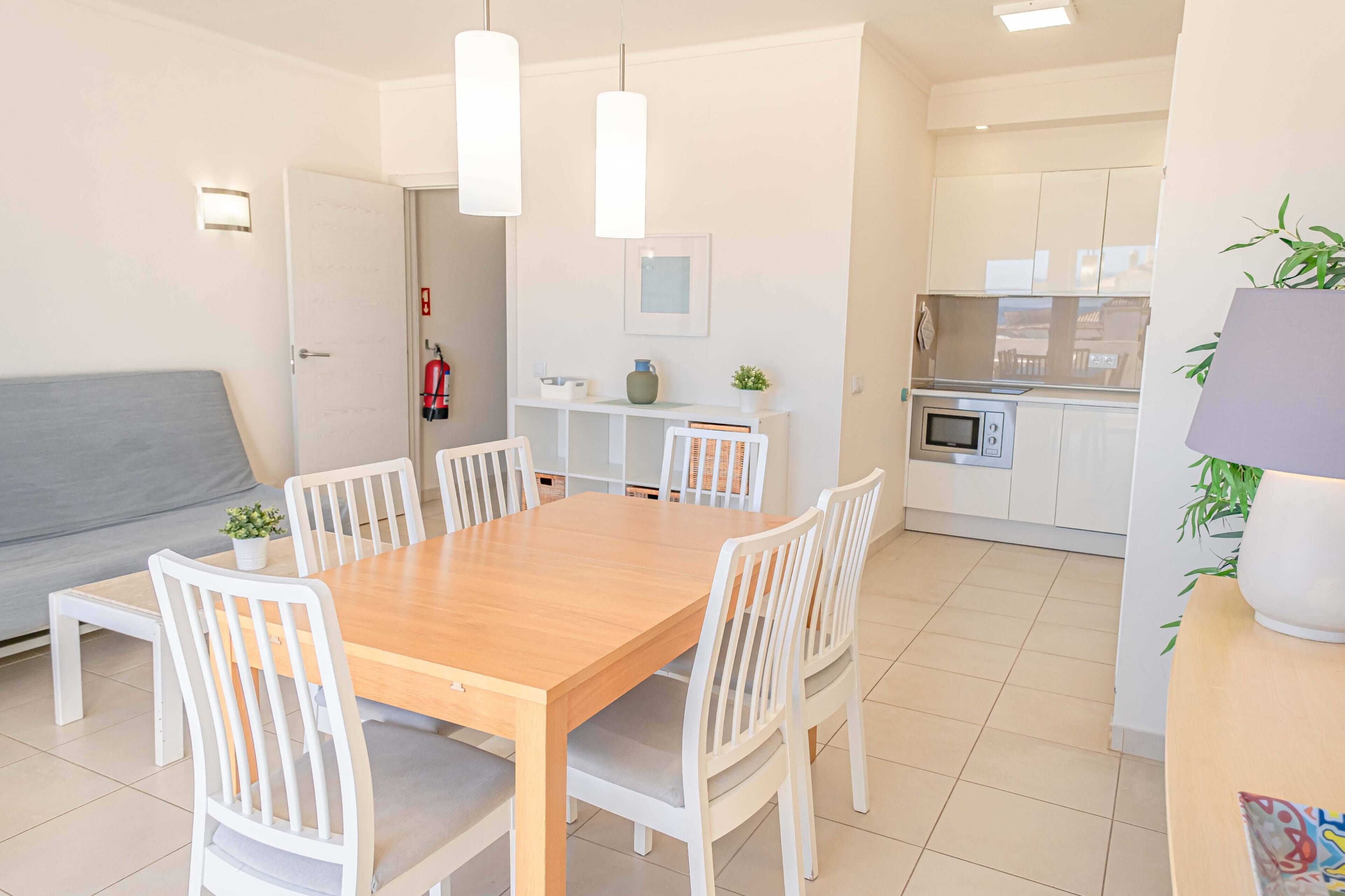 A modern kitchen with white cabinets, a microwave, and a stove. There is a dining area with a wooden table and white chairs. A gray sofa is visible in the background.