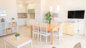 Une cuisine moderne avec des meubles blancs, un coin repas avec une table en bois et des chaises blanches, un téléviseur fixé au mur et une petite plante en pot posée sur le sol.